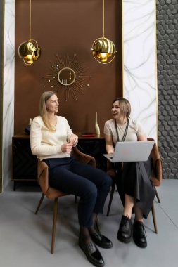 İki kadın şık bir odadaki zarif koltuklarda oturan neşeli bir sohbetin tadını çıkarıyorlar. Tasarımcı dekoruyla çevrili bir dizüstü bilgisayara bakarken fikirleri paylaşıyorlar..