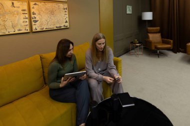 İki kadın şık bir odada sarı bir kanepede oturmuş sohbet ediyorlar. Biri tablet tutarken, diğeri de işbirlikçi bir faaliyeti gösteren küçük bir aygıtı inceliyor..