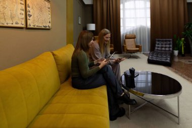 Geniş pencereli ve zarif dekorlu modern bir oturma odasında, iki kadın sarı bir kanepede oturuyor. Mobil cihazlar tutuyorlar ve bir ışık anı paylaşıyorlar, birbirlerine eşlik ederek eğleniyorlar..