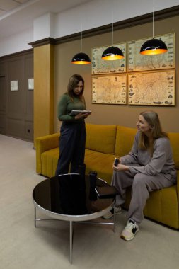 İki kadın, sanatla süslenmiş şık bir odada otururken sohbet ediyorlar. Biri bir defter tutuyor, nişanlı gibi görünüyor, diğeri dikkatlice dinliyor..