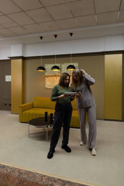 İki arkadaş modern bir odada bir anı paylaşır. Biri bir dergi tutarken diğeri daha yakından bakmak için eğiliyor. Etrafı şık sarı bir kanepe ve yumuşak ışıklandırmayla çevrili..