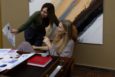 İki meslektaş, çağdaş bir ofiste iş raporları hakkında hararetli bir tartışmaya girişiyor. Biri çizelgelerde yer alırken, diğeri dikkatle dinliyor ve işbirliğini teşvik ediyor..