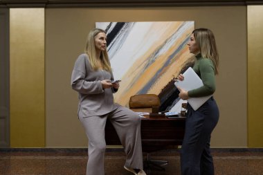 Elinde belgelerle modern bir ofiste sohbet eden iki kadın. Bir kadın masaya yaslanırken, diğeri yakınlarda duruyor, ikisi de çağdaş bir arkaplanda konuşmalarına derinden odaklanmış..