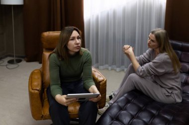 İki kadın, rahat bir oturma odasında derin bir sohbete girişiyor. Bir kadın tablet tutarken, diğeri dikkatle dinliyor, öğleden sonra sıcak bir atmosfer yaratıyor..