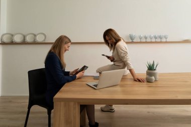 Modern bir ofiste tahta mobilyalarla yan yana oturan iki kadın. Sıradan bir tartışmanın tadını çıkarırken akıllı telefonlarına odaklanıyorlar. Doğal ışık alanı aydınlatır..