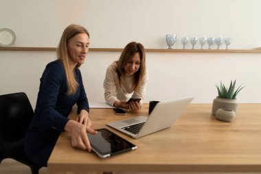 İki kadın, parlak bir çalışma alanındaki ahşap bir masada fikirlerini paylaşırken dizüstü bilgisayar, tablet ve akıllı telefon kullanarak ortak bir tartışmaya giriştiler..
