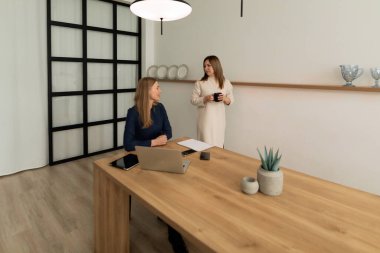 İki kadın doğal ışıkla dolu çağdaş bir ofiste sohbet ediyorlar..