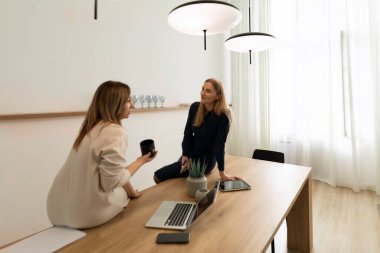 İki kadın şık, ahşap bir masada, parlak bir ofiste canlı bir tartışma içindeler. Bir kadın kahve fincanı tutarken diğeri de yanında dizüstü bilgisayarla masada oturuyor..