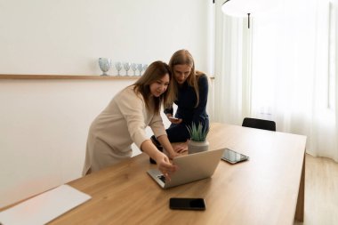 İki kadın bir dizüstü bilgisayara odaklanmış, iyi aydınlatılmış ofis ortamında parlak, havadar dekorlar ve bitkilerle çevrili ahşap bir masada otururken ayrıntıları tartışıyorlar..