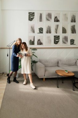 İki kadın modern bir oturma odasında ortak bir faaliyet içindeler. Süslü dekorlar ve bitkilerle çevrili kahve fincanlarını tutarken fikirleri paylaşıyorlar..