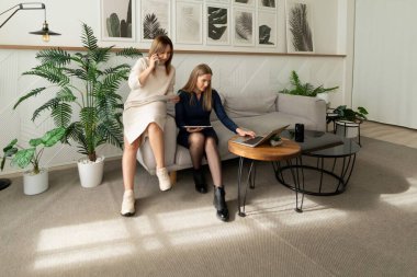 Bitkilerle dolu çağdaş bir yaşam alanında iki kadın işbirliği yapıyor. Bir kadın kanepede otururken diğeri dizüstü bilgisayarla fikir paylaşıyor. Doğal ışık alanı aydınlatır..