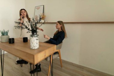 İki kadın kapalı alanda minimalist bir ortamda sohbet ediyorlar. Biri basit bir tahta masada otururken, diğeri ayakta duruyor ve önemli bir şeyi tartışıyor gibi görünüyor..