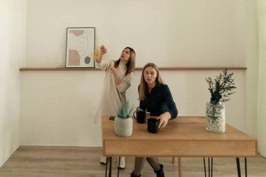 İki kadın minimalist bir odada ahşap bir masada otururken, biri bir objeyi incelerken diğeri de kahve fincanından yudumlayarak sıcak ve rahat bir atmosfer yaratıyor..