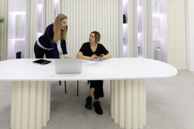 Zarif ve çağdaş bir ofis ortamında, iki kadın bir dizüstü bilgisayar ve belgelerle şık bir masada verimli bir tartışmaya girişiyor. Atmosfer odaklı ve dinamik..