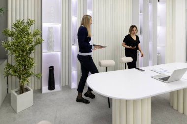 İki kadın şık ve modern bir ofis içinde ortak bir faaliyet içindeler. Bir kadın bir tepsi taşırken diğeri fikirleri tartışıyor gibi görünüyor..