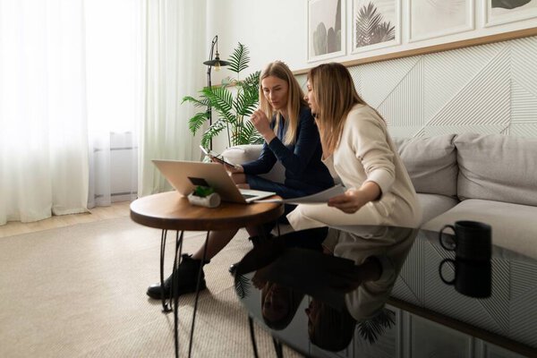 Две женщины сидят на удобном диване, участвуя в обсуждении во время просмотра документов и с помощью ноутбука, в окружении стильной обстановки гостиной.