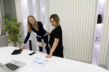 Çağdaş bir ofiste iki kadın ortak bir toplantıya katılıyor. Biri bir belgeyi incelerken, diğeri önemli verileri vurgulayarak dijital bir cihaza doğru hareket eder..