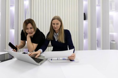 İki kadın şık, aydınlık bir çalışma alanında ortak bir tartışmaya giriştiler. Biri belgeleri incelerken, diğeri de akıllı telefondan izleyip takım çalışmasını destekliyor..