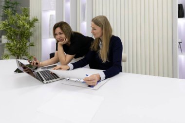 İki kadın bir ofisteki şık bir masada birlikte çalışıyor. Biri bir dizüstü bilgisayara odaklanırken, diğeri de gün ortası ışığında verimli bir tartışmayla meşgul olan belgeleri inceliyor..