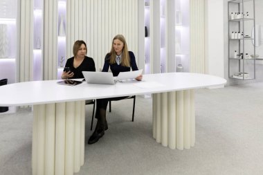 İki kadın zarif bir dekor ve yumuşak ışıklandırmayla çevrili çağdaş bir ofis ortamında, gösterişli beyaz bir masada otururken sohbet ediyorlar..