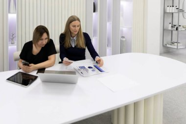 İki kadın şık, modern bir masada iş görüşmesi yapıyor. Biri evrakları incelerken diğeri de telefonuna bakıyor. Etrafı minimalist ofis zeminiyle çevrili..