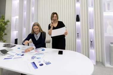 İki meslektaş, iyi aydınlatılmış bir ofis alanında proje değerlendirmesi sırasında heyecanlarını dile getirdiler. Biri raporları analiz ederken, diğeri coşkuyla hareket ediyor, takım çalışması ve başarı sergiliyor..