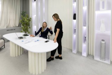 İki kadın şık, modern bir ofis masasında tartışırlar. Biri dizüstü bilgisayara dikkatle bakarken diğeri ayakta duruyor ve şık bir arkaplandaki fikirlere katkıda bulunuyor..