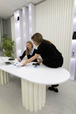 İki profesyonel, şık bir ofis masasında ortak bir tartışmaya girişiyor, iyi aydınlatılmış bir çalışma alanındaki dizüstü bilgisayardaki belge ve tasarımları gözden geçiriyorlar..