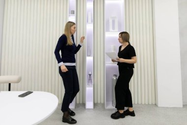 İki kadın şık bir odada şık bir dekorla dururken fikirleri tartışıyor. Bir kadın kağıtları tutarken diğeri jestler yaparak aralarında dinamik bir etkileşim yaratıyor..