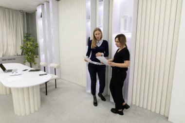 İki kadın, çağdaş bir ofis alanındaki proje belgeleri hakkında sohbet ediyorlar. Oda, nötr renkleri ve modern mobilyaları olan gösterişli bir tasarıma sahip..