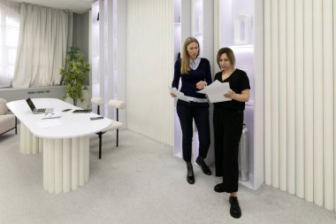 İki kadın çağdaş bir ofiste dururken proje materyallerini tartışıyor. Kağıtları gözden geçirir ve zarif dekorlarla dolu aydınlık bir ortamda görüşlerini paylaşırlar..