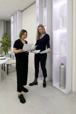 Çağdaş bir ofis ortamında, parlak tasarım unsurları ve sakinleştirici renklerin yer aldığı belgeleri incelerken, iki kadın işbirliği içinde tartışıyorlar..