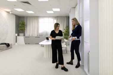 Çağdaş bir ofis alanında iki kadın iş birliği yapıyor. Biri belgeleri sunarken, diğeri onları inceliyor, takım çalışmalarını ve verimli bir ortamda çalışmalarını gösteriyor..