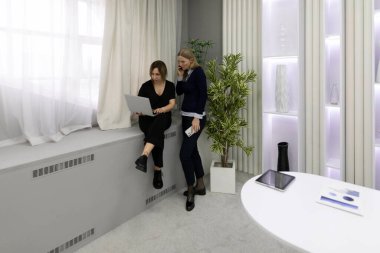 Bir kadın pencerenin kenarında dizüstü bilgisayarla otururken, diğeri de yanında durmuş sohbet ediyor. Modern ofis parlak ve minimalist bitkiler ve yumuşak ışıklandırma ile.