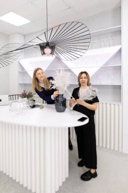 Çağdaş bir dükkanda iki kadın çiçek aranjmanı yapıyor. Şık dekorasyon ve minimalist tasarım davetkar atmosferi arttırır. İşlerine odaklanmış ve meşguller..