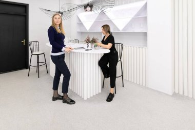 İki kadın, eşsiz bir bar tarzı masa ve zarif bir dekorun yer aldığı çağdaş bir beyaz çalışma alanında sohbet ediyorlar. Çevre minimalizmi ve karmaşıklığı vurguluyor.