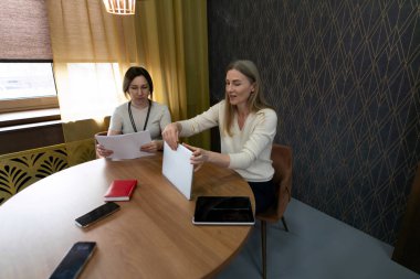 Çağdaş bir ofis ortamında, yuvarlak tahta bir masada oturan iki kadın, belgelerini değiş tokuş ediyor ve teknolojiyi kullanırken işle ilgili konuları tartışıyorlar..