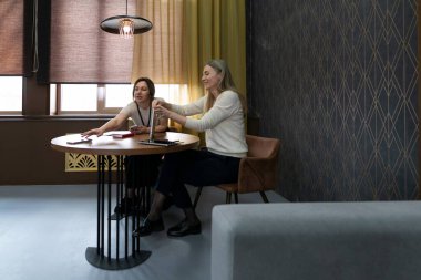 İki kadın şık bir ofiste, yuvarlak bir masada tartışırlar. Belgelere bakıyorlar ve yumuşak ışıklandırmalı rahat bir atmosferde fikirleri paylaşıyorlar..