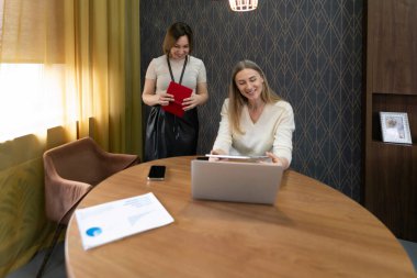 İki profesyonel, yuvarlak ahşap bir masada dizüstü bilgisayarın üzerinde tartışır. Bir kişi elinde bir defter tutarken gülümsüyor ve modern bir ofisteki ortak çalışma ortamını vurguluyor..