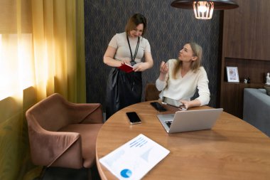 Günlük kıyafetli iki kadın, tahta bir masanın etrafında dizüstü bilgisayar ve belgelerle etkileşim halindeler. Sıcak ışık perdelerden süzülerek çağdaş bir ofiste üretken bir atmosfer yaratıyor..