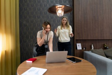 İki kadın şık bir ofiste, biri telefonda diğeri ise yuvarlak bir masada dizüstü bilgisayar kullanarak bir projede işbirliği yaparken el kol hareketi yapıyor. Onları çevreleyen hoş bir atmosfer var..