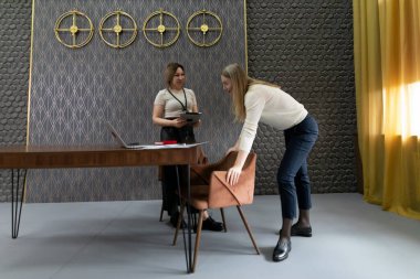 İki kadın şık bir ofiste masanın yanında dikilirken sohbet ediyorlar. Bir kadın sandalyeye yaslanırken, diğeri tablet tutuyor, düşüncelerini paylaşıyor..