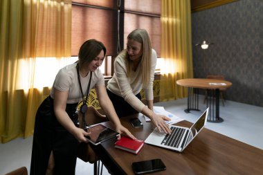Kadınlar, günışığıyla dolu çağdaş bir ofis ortamındaki ahşap bir masa üzerindeki dizüstü bilgisayar ve tabletler gibi cihazları kullanarak belgeleri incelerken tartışmalara giriyorlar..