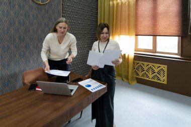 Çağdaş bir ofiste çalışan iki kadın, bir masada dizüstü bilgisayar ve çizelge ile otururken belgeleri inceleyip proje detaylarını tartışıyor..