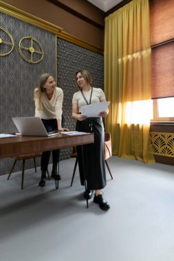 İki profesyonel kadın, dekoratif unsurlarla süslü bir ofiste işbirliği yapıyor ve belgeleri paylaşıyorlar. Geniş pencereler doğal ışık girmesini sağlıyor ve verimli bir atmosfer yaratıyor..