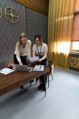 İki kadın, modern bir ofis masasında verimli bir tartışma içinde, bilgisayardaki belgeleri incelerken, büyük pencerelerden akan doğal ışığın keyfini çıkarıyorlar..