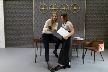 Çağdaş bir çalışma alanında iki kadın ortak bir tartışmaya giriyorlar. Biri akıllı telefondan içeriğini gösterirken, diğeri tablet taşıyan bir masaya eğilip şık bir arka planla çevrili..