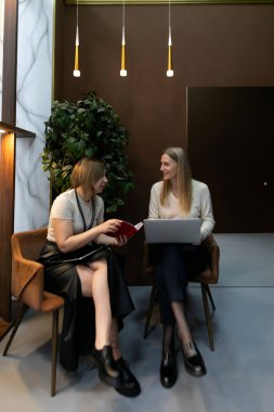 İki kadın şık koltuklarda oturuyor, biri diğerine bir kitap gösteriyor, dizüstü bilgisayarda çalışıyor, çağdaş dekorlar ve sıcak aydınlatmalarla çevrili..