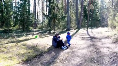 Mavi giyinmiş bir çocuk yakınlarda oynarken bir yetişkin dingin bir ormanda bir ağaca yaslanıyor. Güneş ışığı ağaçların arasından süzülüp rahatlatıcı bir atmosfer yaratır..