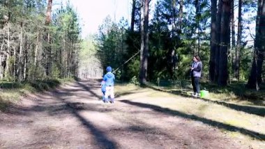 Küçük bir çocuk, uzun ağaçlarla kaplı bir ormanda scooter sürerken bir ebeveyn onu izlerken mutlu bir şekilde seyrediyor. Açık hava aktiviteleri için güneşli bir gün..